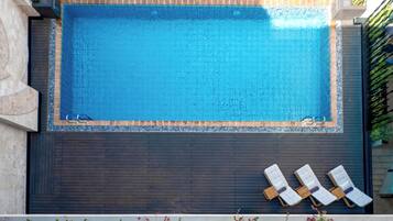 Una piscina rectangular con cubierta de madera oscura y tres sillas reclinables blancas.