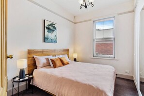 Een netjes ingerichte slaapkamer met een groot bed, een nachtkastje met een lamp, een raam met jaloezieën en een schilderij aan de muur.