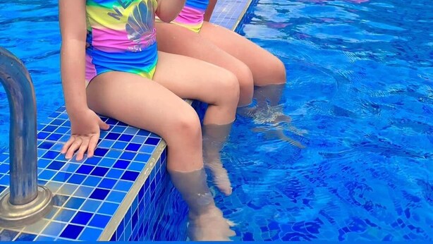 Dos niños sentados al borde de una piscina, con un flamenco inflable rosa al fondo.