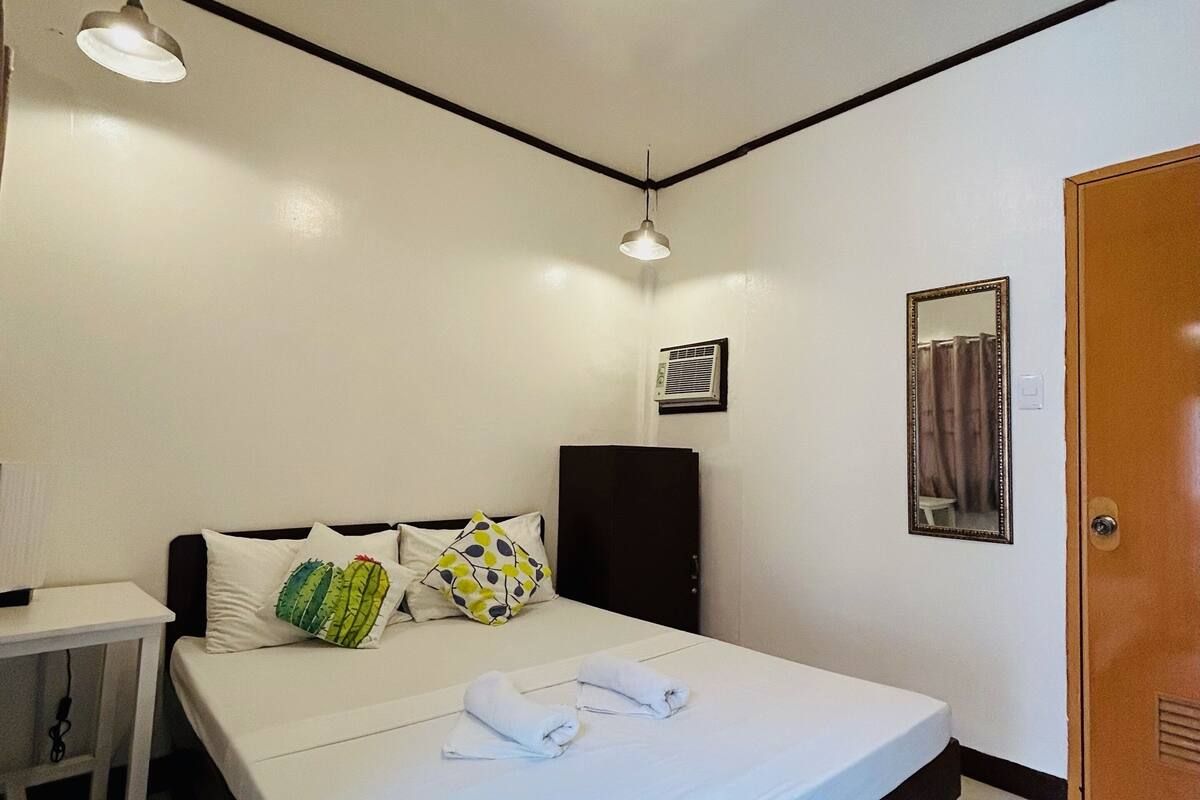 A hotel room with a bed, bedside table, lamp, air conditioning unit, and a door.