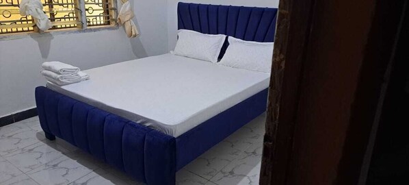 Ein ordentlich bezogenes Bett mit weißer Bettwäsche und einem blauen Kopfteil in einem Raum mit Fliesenboden.