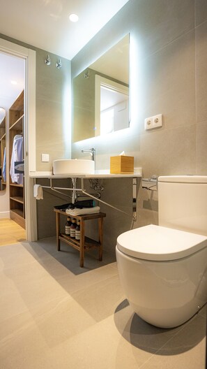 A modern bathroom with a large toilet, a sink, and a mirror. There is a wooden stool with bottles and a towel. - Casa Pince (Barcelona)