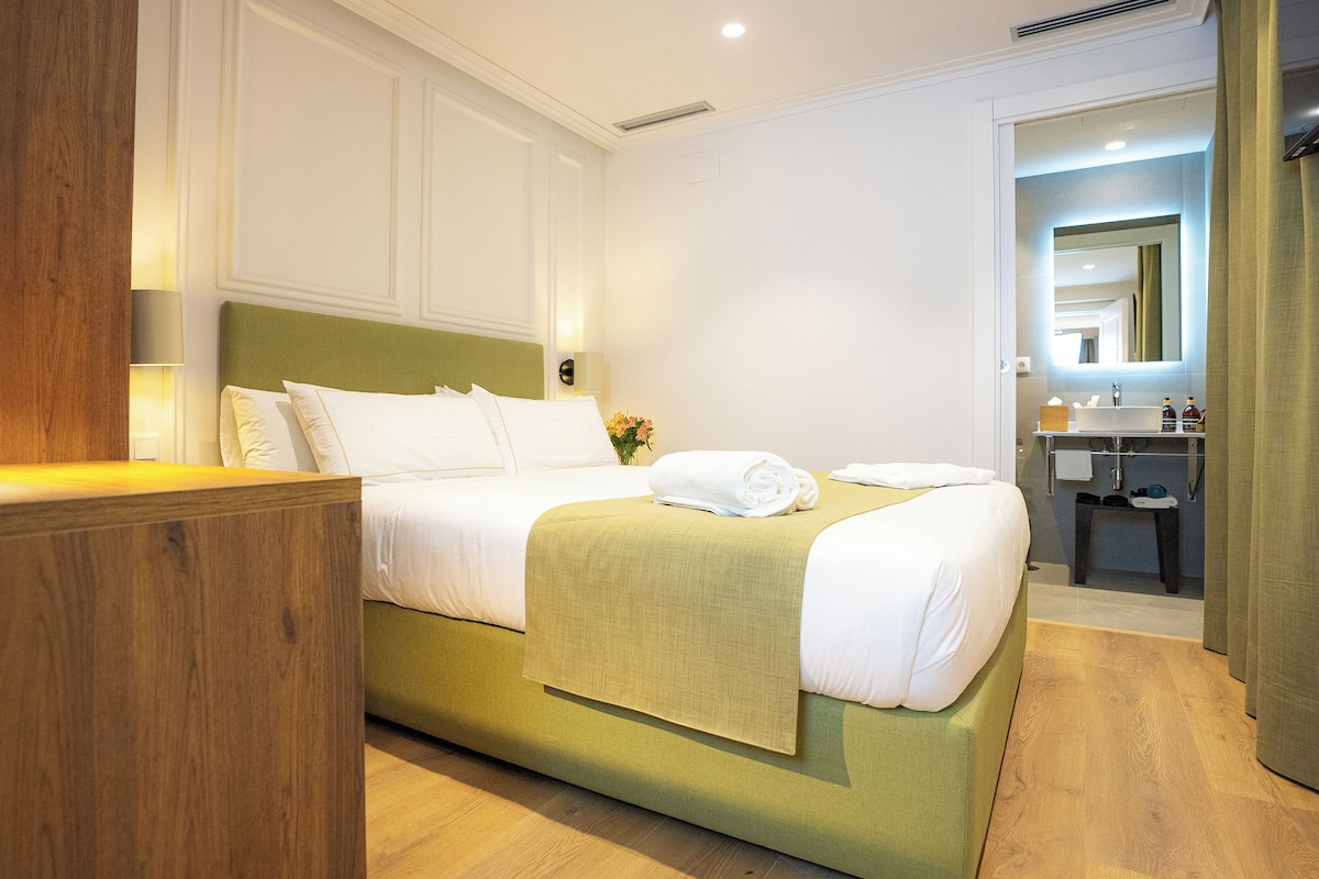 A hotel room with a bed, a bedside table, a mirror, and a bathroom visible through an open door.