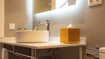 Un baño moderno con un espejo grande, un lavabo y un inodoro. Hay una pequeña estantería debajo del lavabo con botellas.