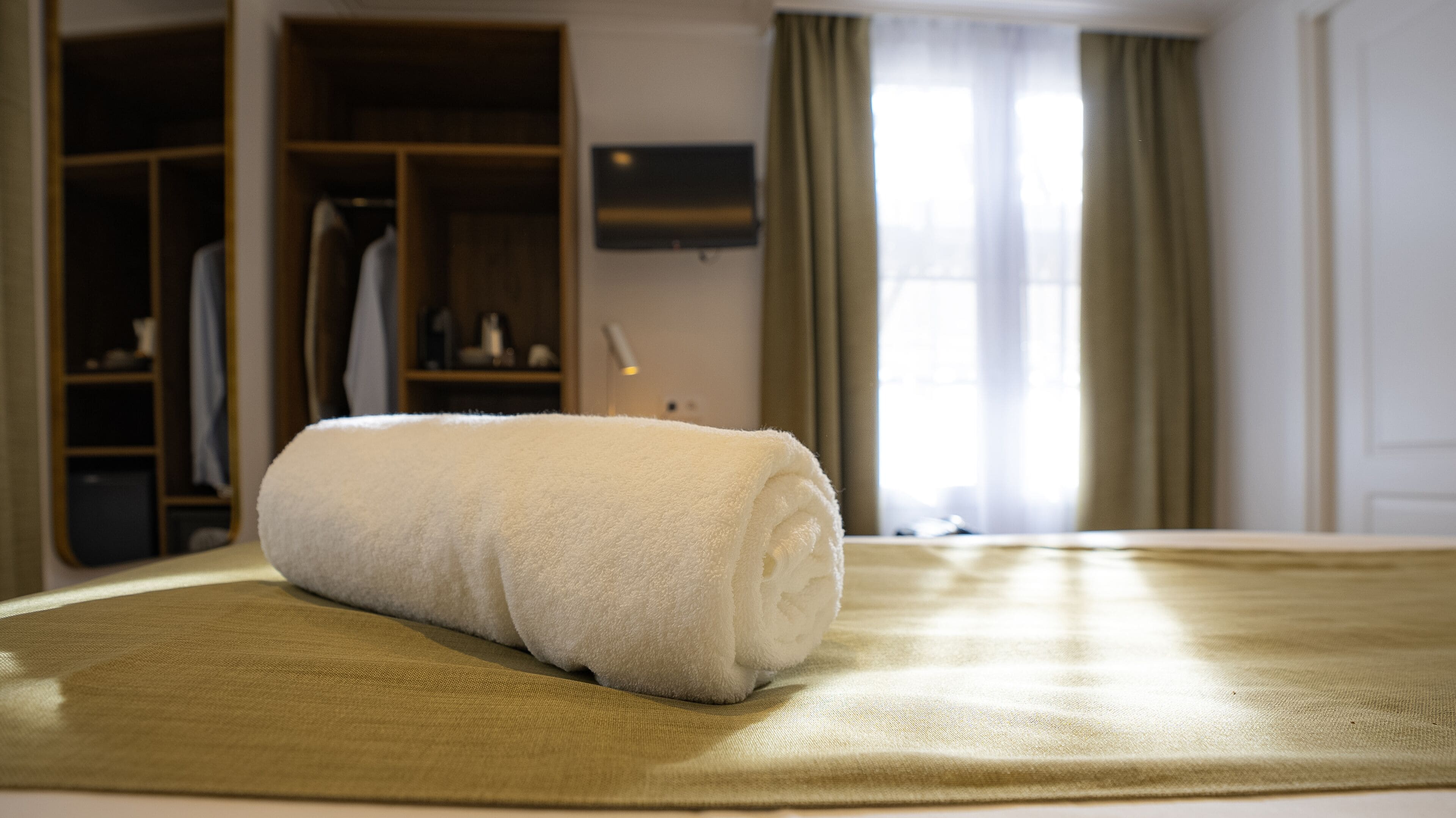 A neatly made bed with a rolled white towel, a wooden wardrobe, and a television on the wall.