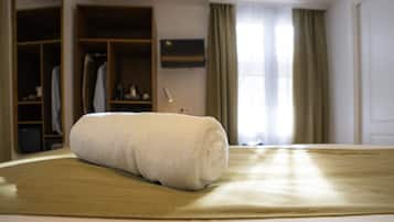 A neatly made bed with a rolled white towel, a wooden wardrobe, and a television on the wall.
