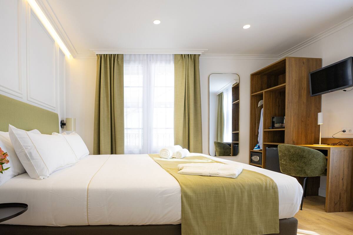A hotel room with a large bed, a desk, a chair, a TV, and a wardrobe.