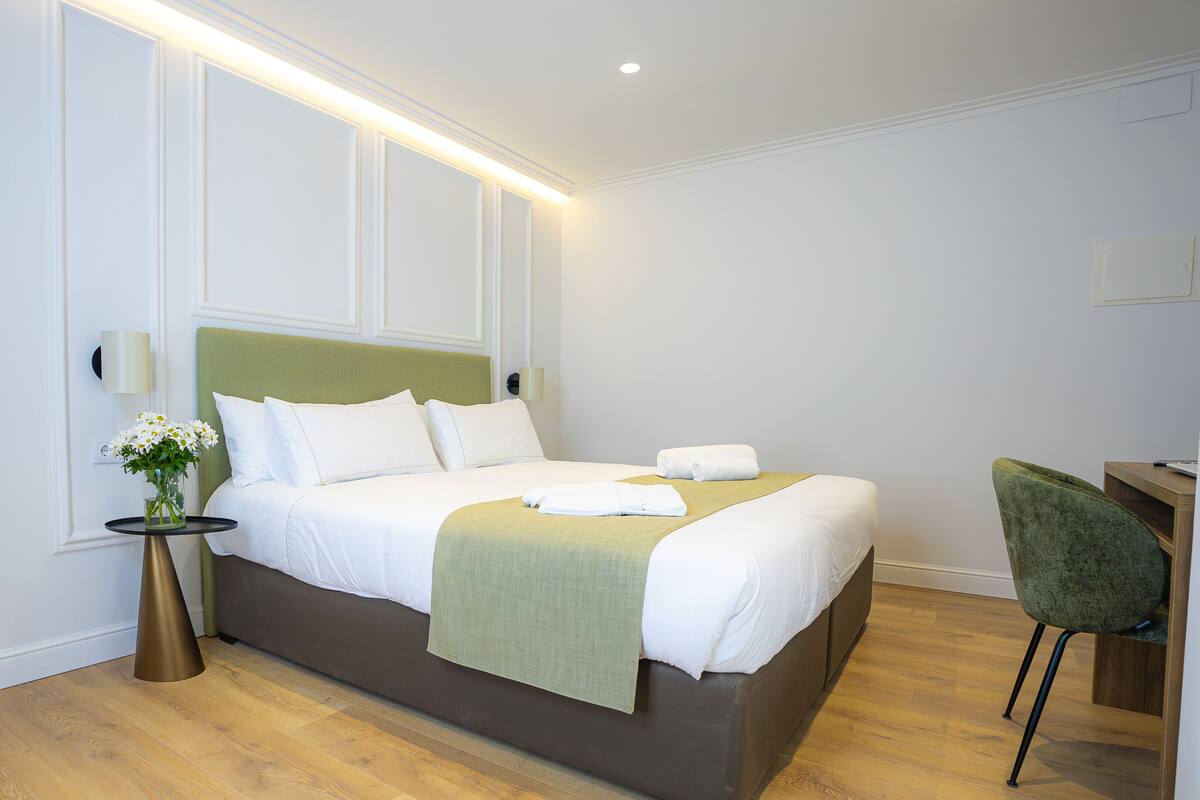 A neatly made bed with white linens, a green throw blanket, and a vase of flowers on a round table beside it.