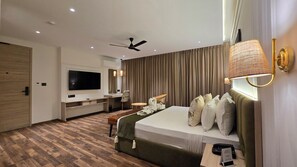 Habitación de hotel moderna con televisor de pantalla plana, ventilador de techo, cama con cabecero verde, mesita de noche y ventana con cortinas.