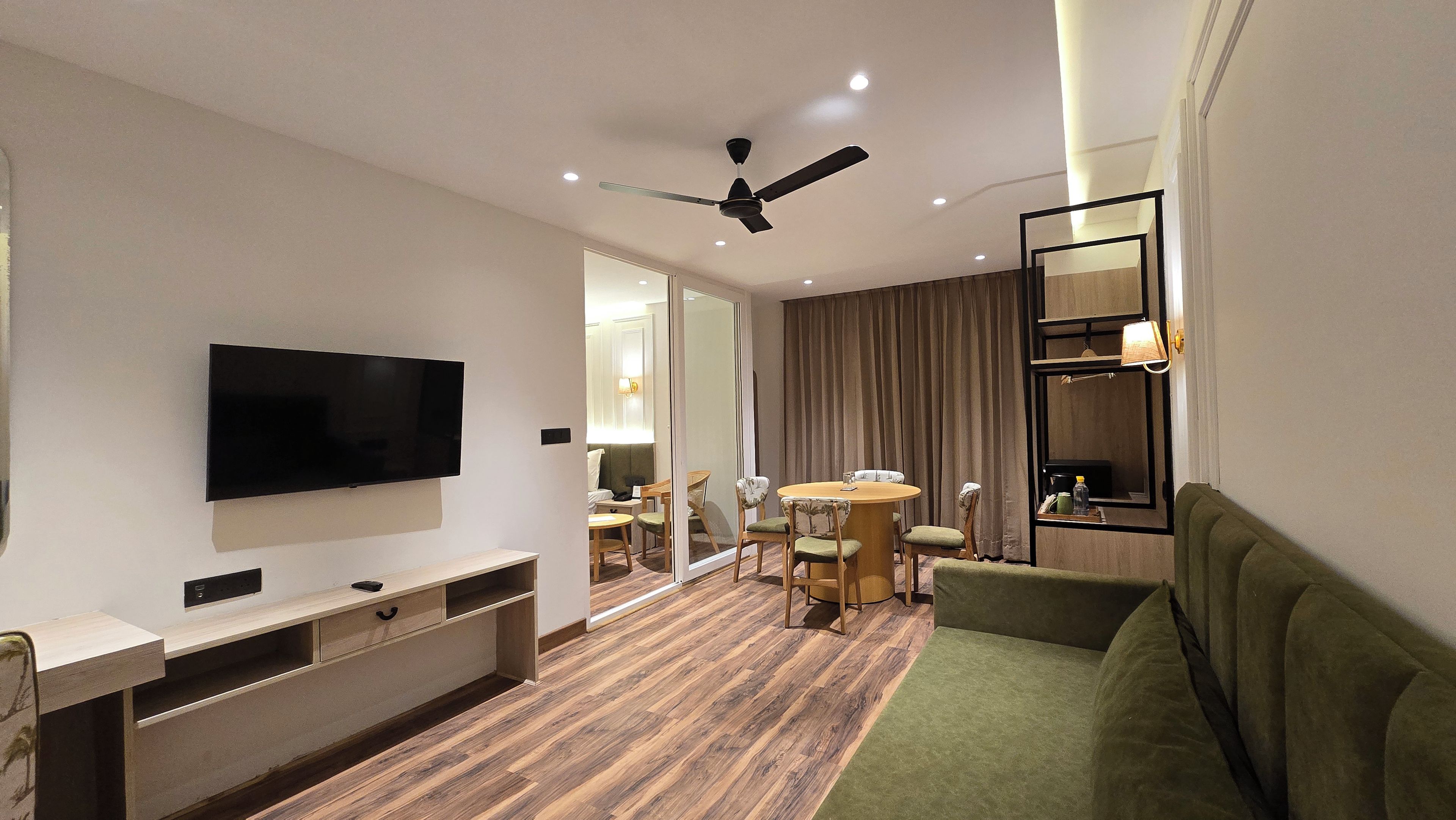 Habitación de hotel con televisor de pantalla plana, ventilador de techo, zona de comedor con mesa y sillas, y sofá verde.