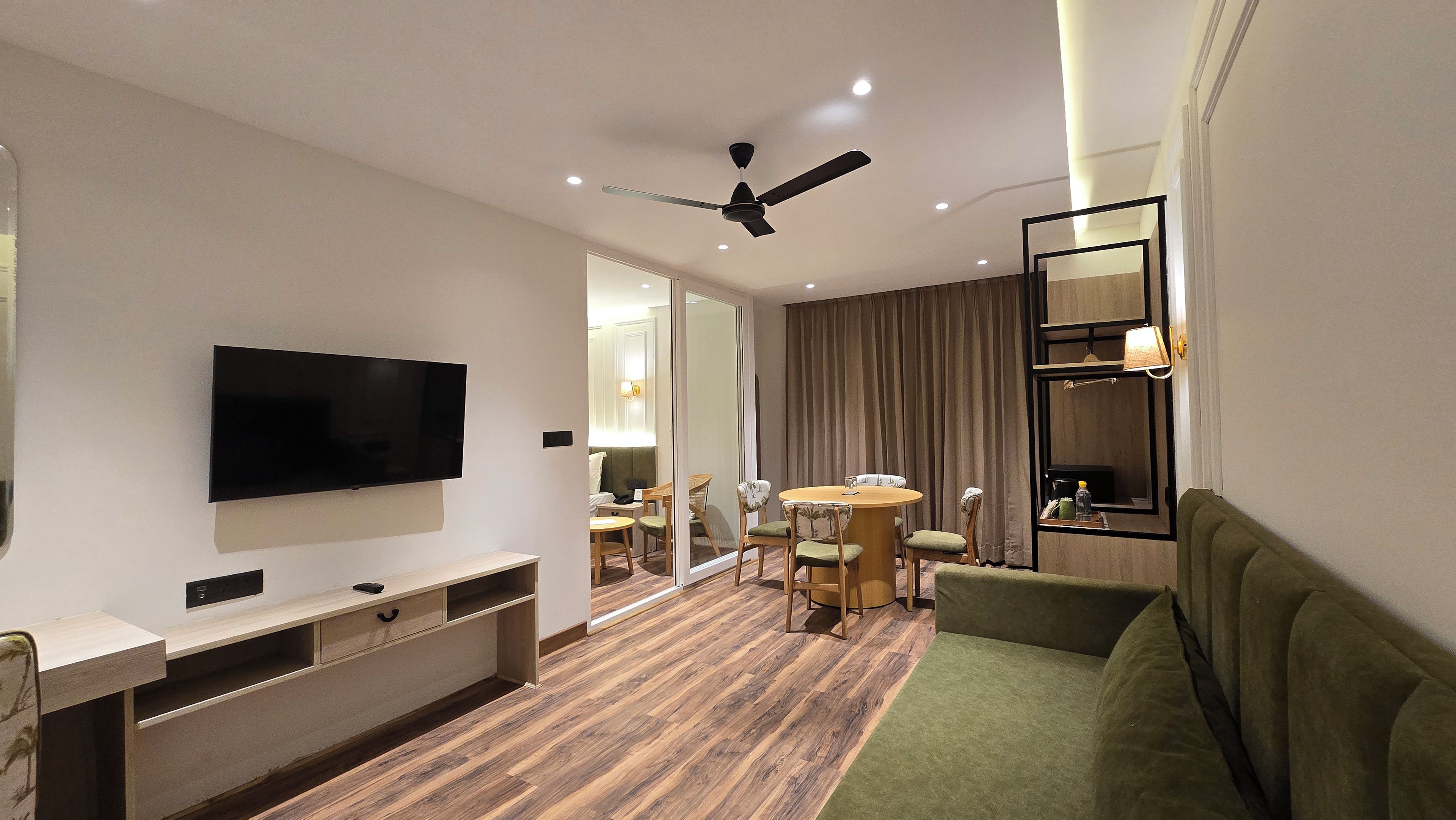 Habitación de hotel con televisor de pantalla plana, ventilador de techo, comedor con mesa y sillas, y un sofá verde.