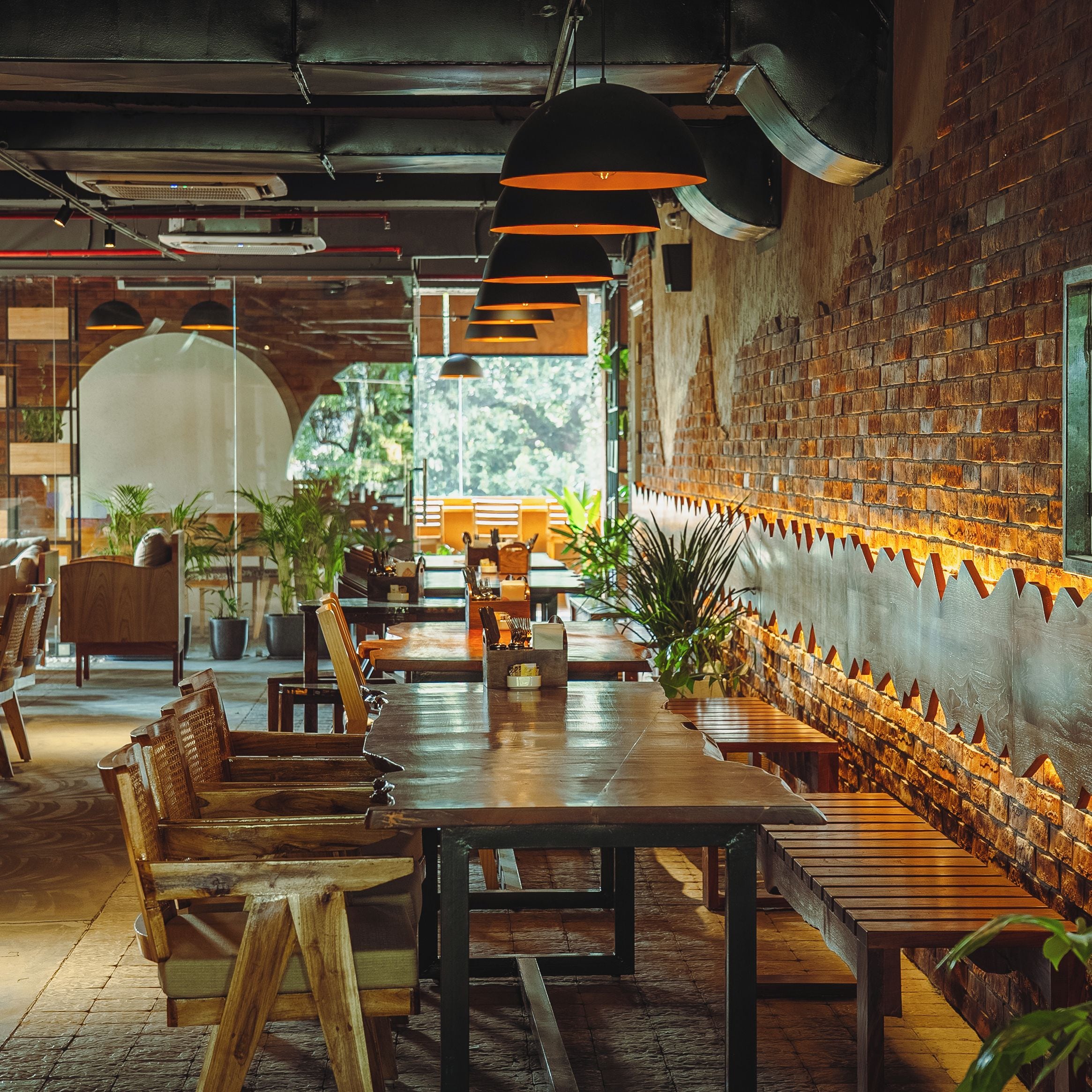 A restaurant interior with wooden tables and chairs, brick walls, and hanging pendant lights.