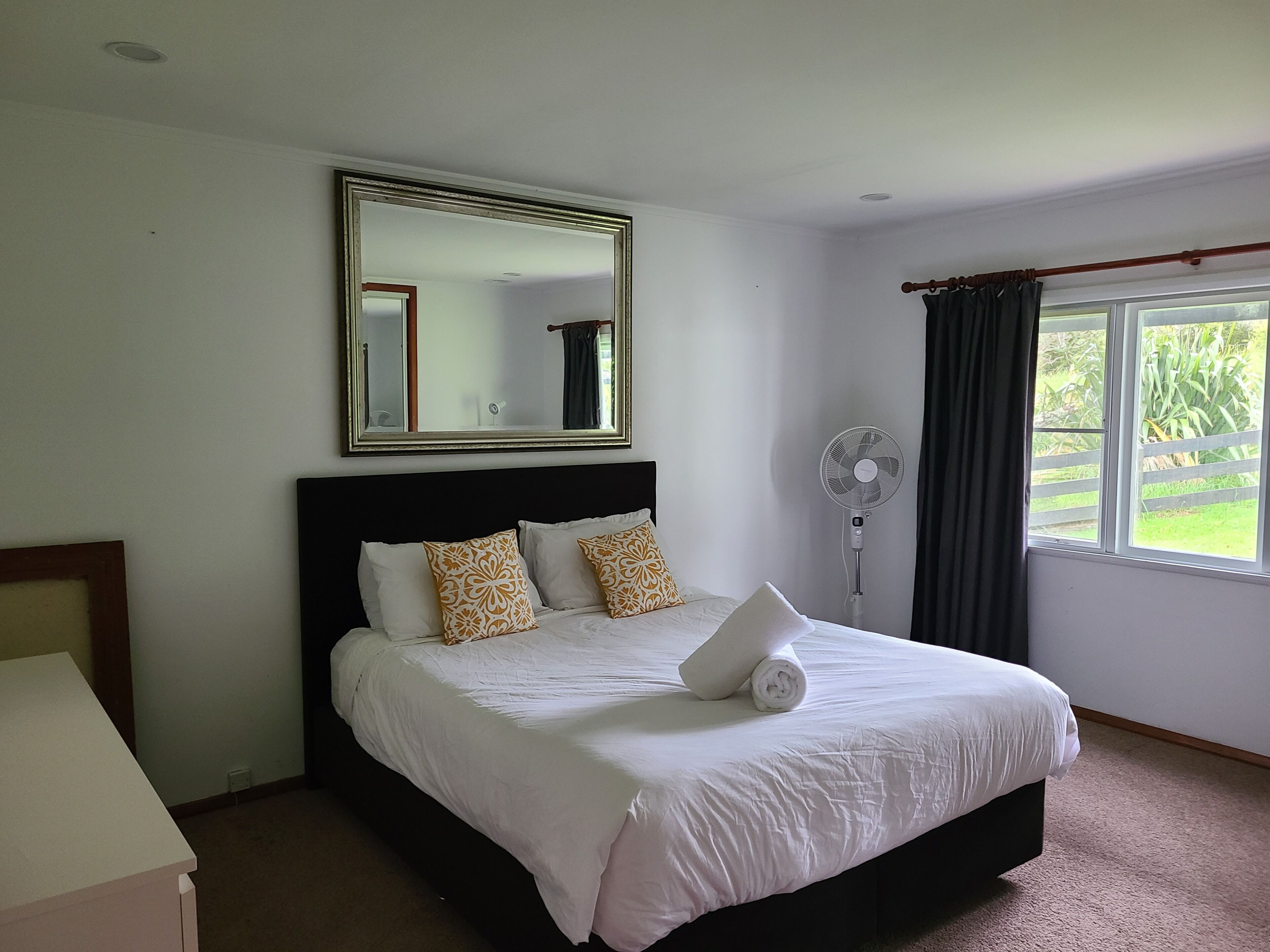 A bedroom with a bed, a standing fan, a mirror, and a window with curtains.