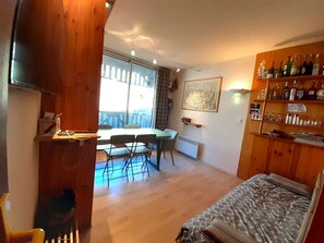Une pièce comprenant un lit, une table à manger avec des chaises, un radiateur, une étagère en bois avec des bouteilles et une grande fenêtre.