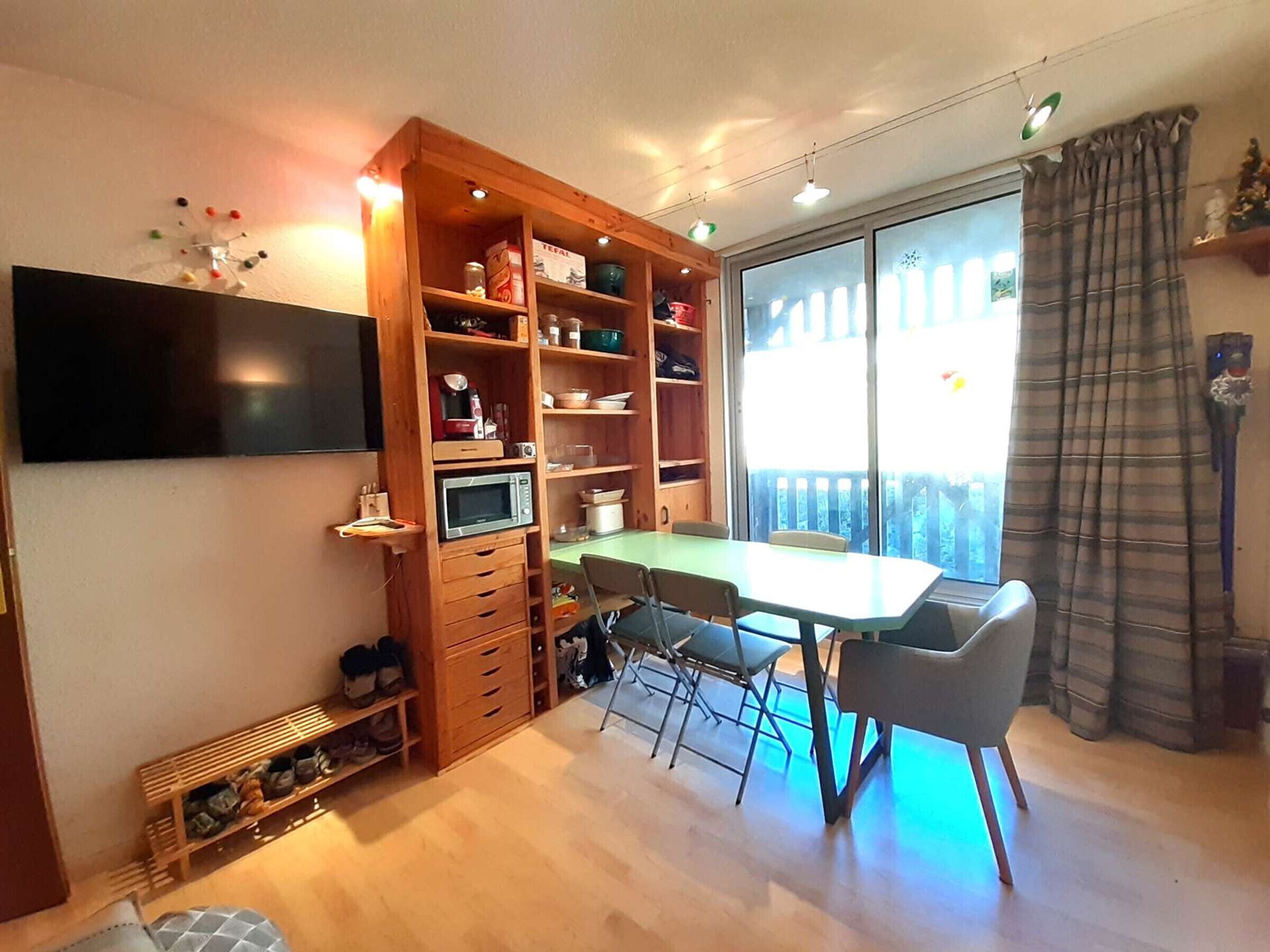 Ett rum med ett matbord, stolar, en tv som är monterad på väggen, ett träskåp med lådor och ett fönster med utsikt.