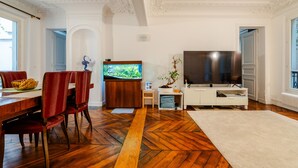 Ein Wohnzimmer mit Holzdielen im Fischgrätmuster, einem Essbereich mit Holztisch und roten Stühlen, einem Flachbildfernseher und einem weißen Sofa.
