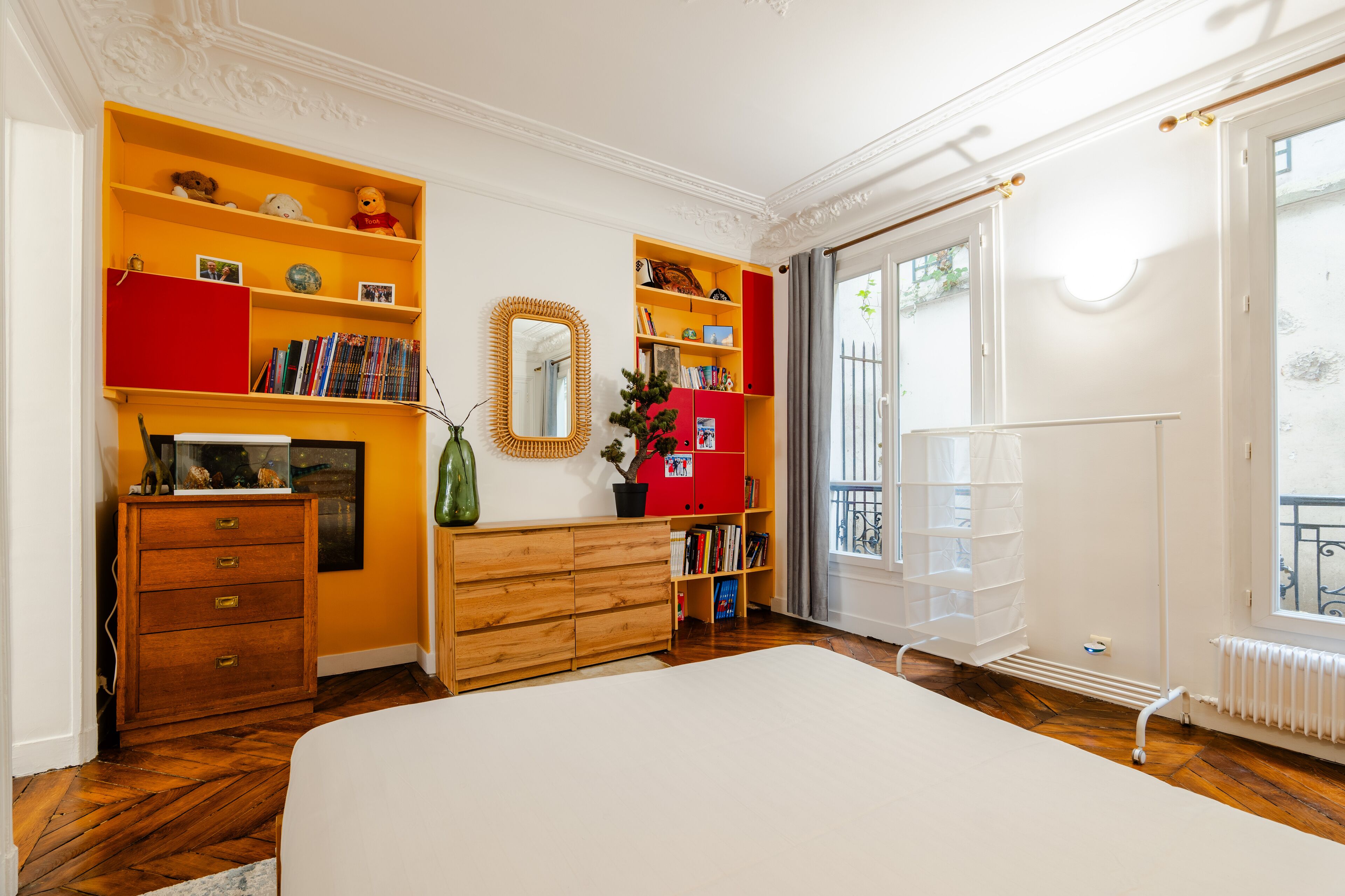 Ein Schlafzimmer mit einem großen Bett, einer Holzkommode, roten und gelben Regaleinheiten, einem Spiegel und einem Fenster mit Blick nach außen.