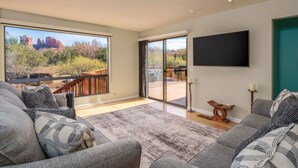 A spacious living room with a large sliding glass door leading to a balcony, a flat-screen TV mounted on the wall, and a cozy seating area with patterned cushions. - Cathedral Views | Large home with two living spaces and kitchens (Sedona)