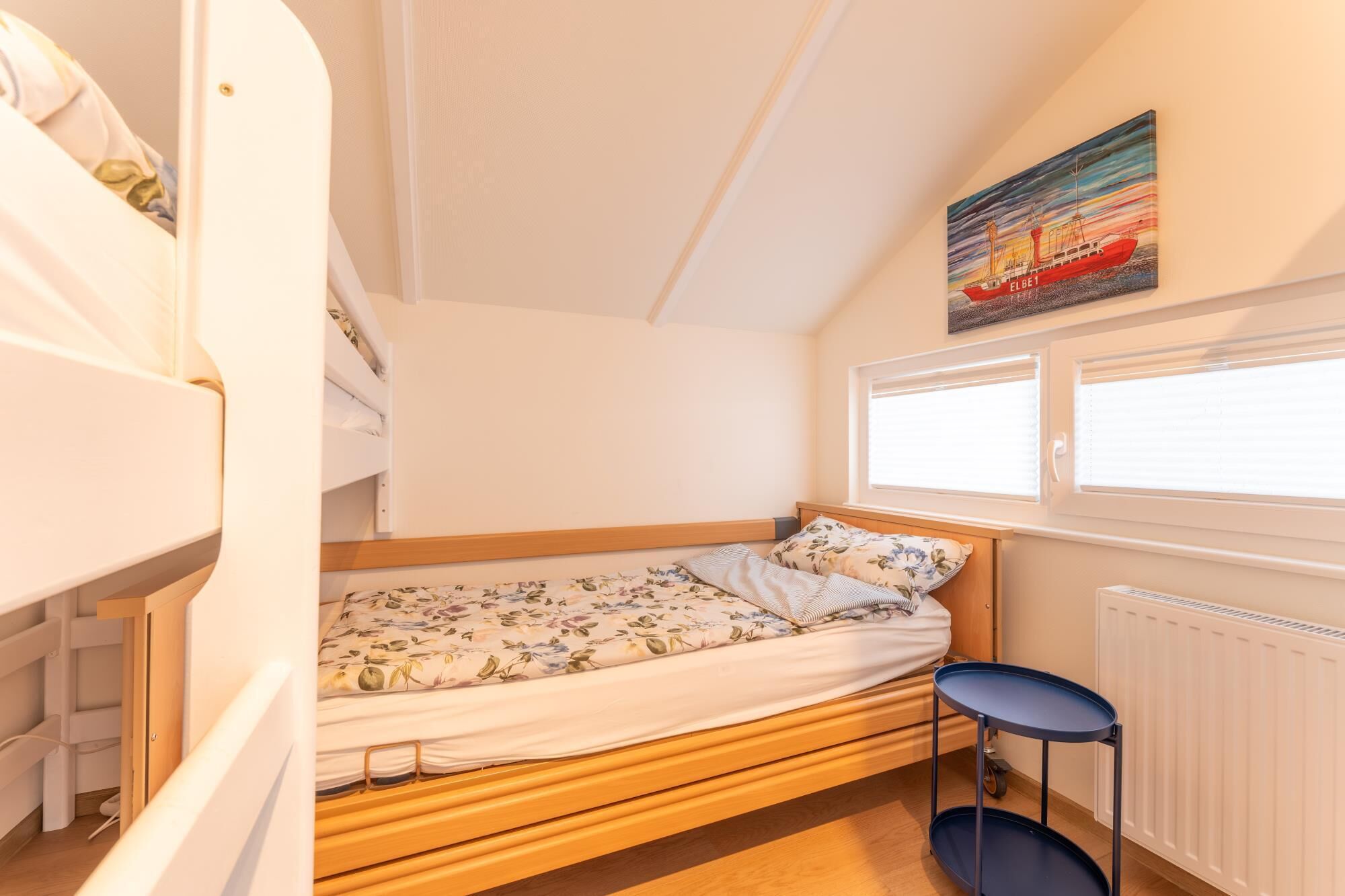 Quarto pequeno e compacto com uma cama de solteiro, uma cama individual com roupa de cama floral, uma mesinha e uma janela com persianas.