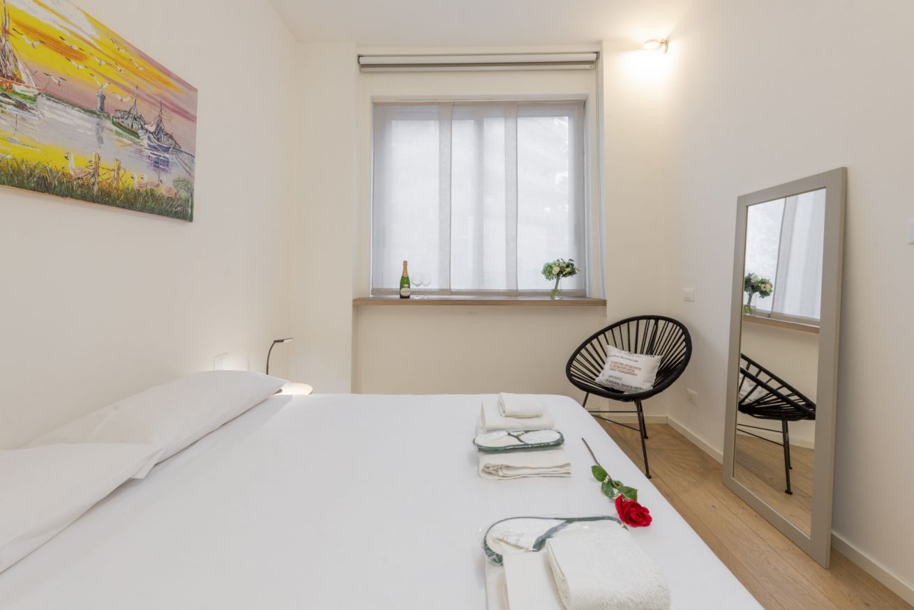 Ein Hotelzimmer mit Bett, einem Fenster mit durchsichtigen Vorhängen, einem Sessel, einem Spiegel und einem Gemälde an der Wand.