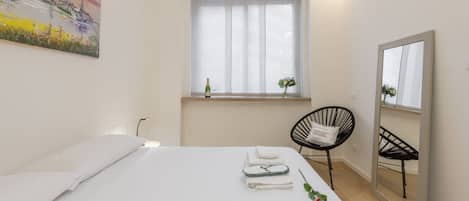 Une chambre d’hôtel avec un lit, une fenêtre avec des rideaux légers, une chaise, un miroir et un tableau accroché au mur.