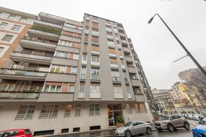 Un immeuble résidentiel à plusieurs étages, doté de balcons, avec une entrée bien visible et des voitures garées dans la rue.