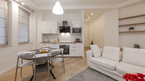 A modern kitchen with white cabinets, a dining table with chairs, and a white sofa. - BnButler - Longhena, 7 (Milano)
