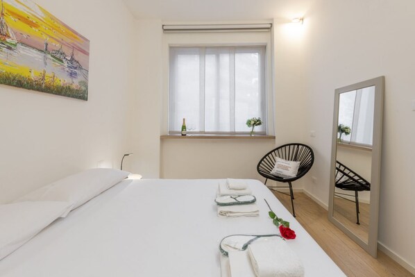 A hotel room with a bed, a window with sheer curtains, a chair, a mirror, and a painting on the wall.