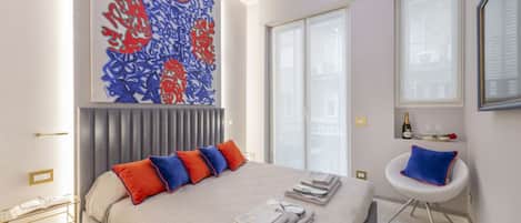 Una habitación de hotel moderna con una cama grande, un elemento decorativo en la pared, una ventana con persianas, una mesita con una botella y vasos, y una silla con cojín.