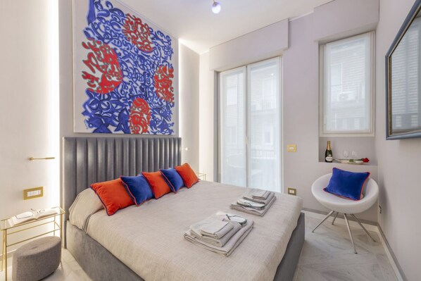 A modern hotel room with a large bed, a decorative wall piece, a window with blinds, a small table with a bottle and glasses, and a chair with a cushion.