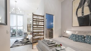 A modern bedroom with a bed, a view of the sky, a balcony, and a large painting.
