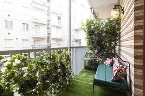 Un balcon avec un revêtement de gazon synthétique, un banc en métal avec des coussins et une plante en pot.