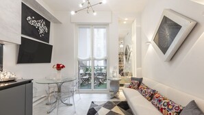A modern living room with a sofa, a glass table, and chairs, a kitchen area with a counter and cabinets, and artwork on the walls.