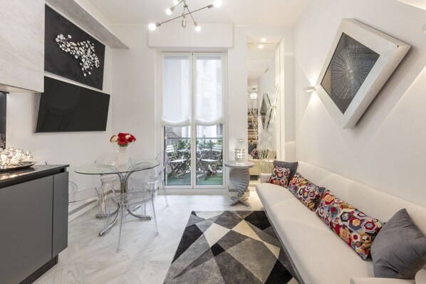A modern living room with a sofa, a glass table, and chairs, a kitchen area with a counter and cabinets, and artwork on the walls.