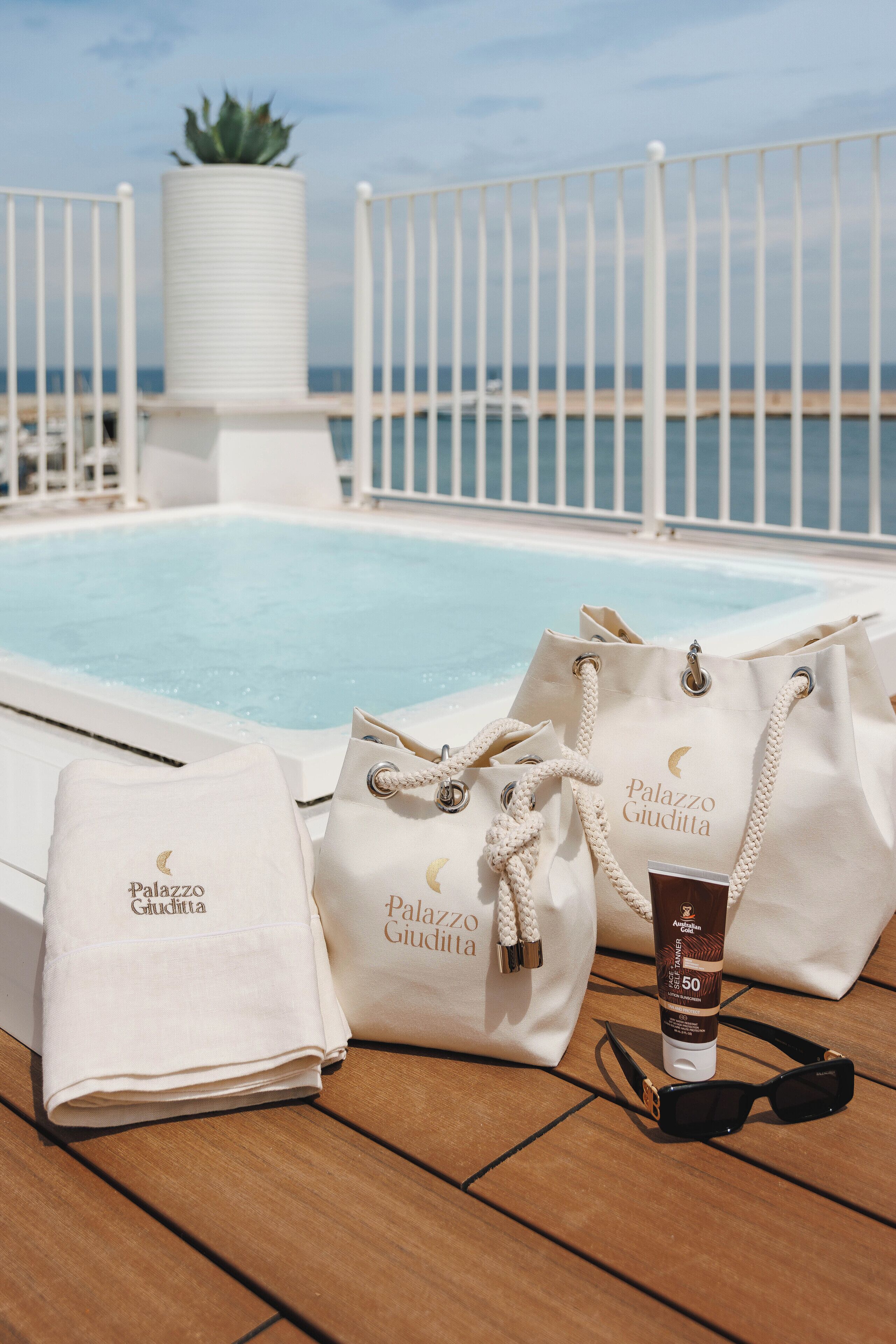 Un espace aménagé au bord de la piscine, avec une clôture blanche, un pot de fleurs et une piscine. On y trouve deux sacs blancs portant la marque « Palazzo Giuditta », une bouteille de crème solaire et une paire de lunettes de soleil, le tout posé sur la terrasse en bois.