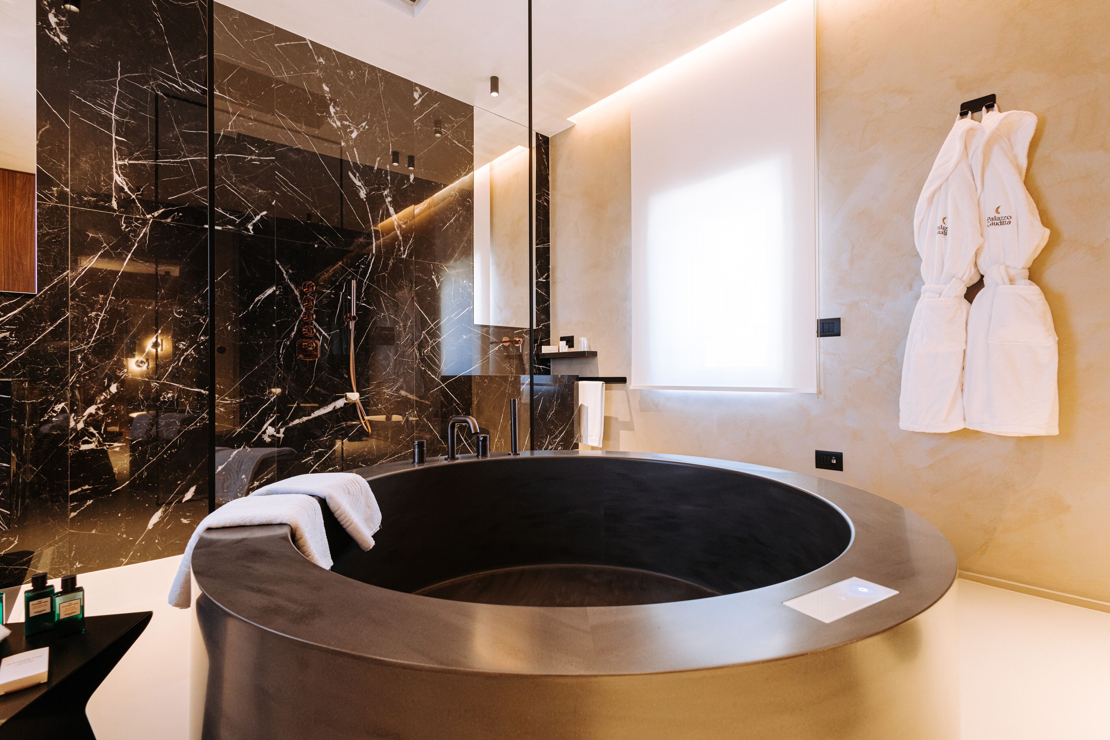 A modern bathroom with a marble wall, a freestanding bathtub, and two white robes hanging on the wall.