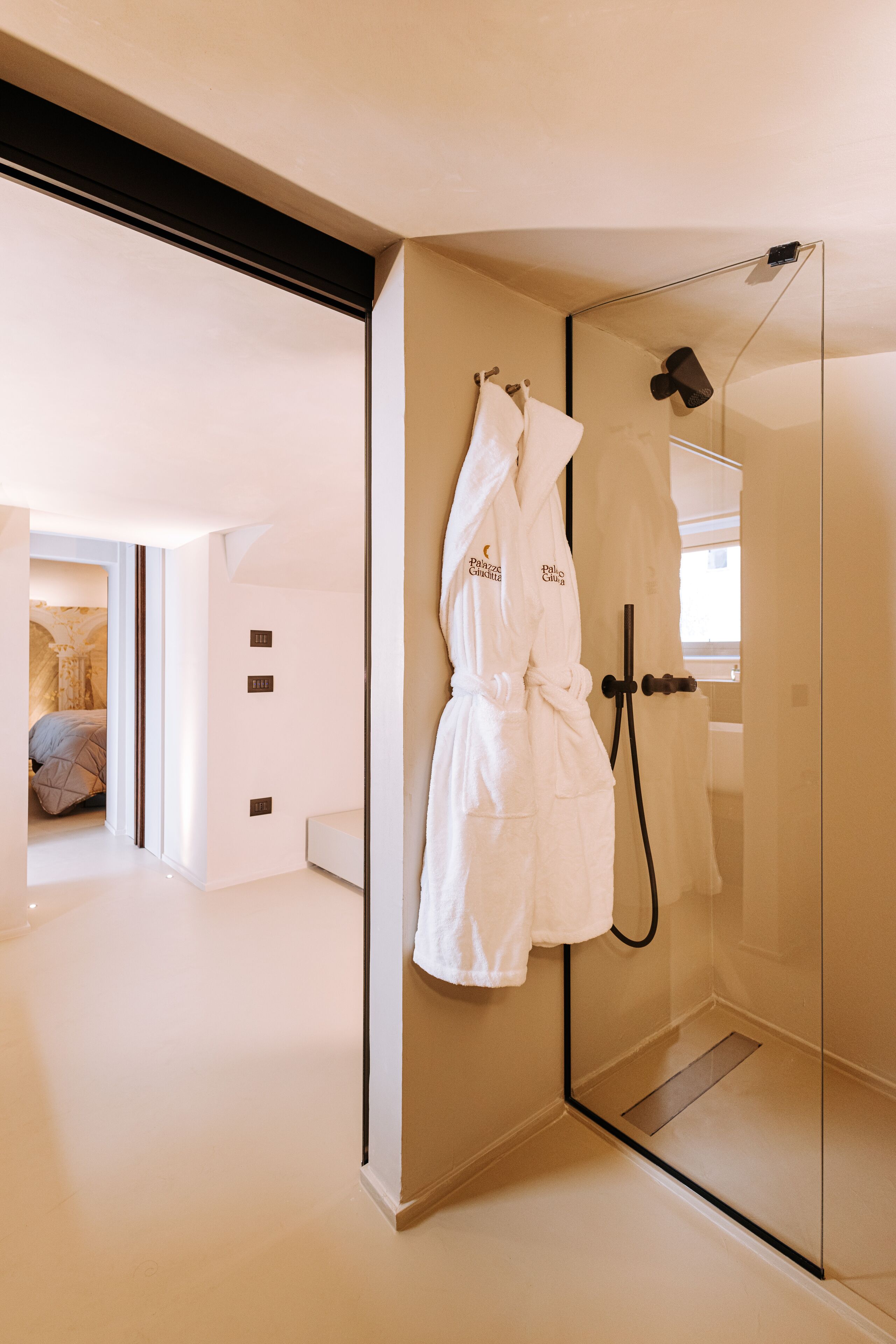 A modern bathroom with a glass shower enclosure, a white robe hanging on the wall, and a visible bed in the adjacent room.