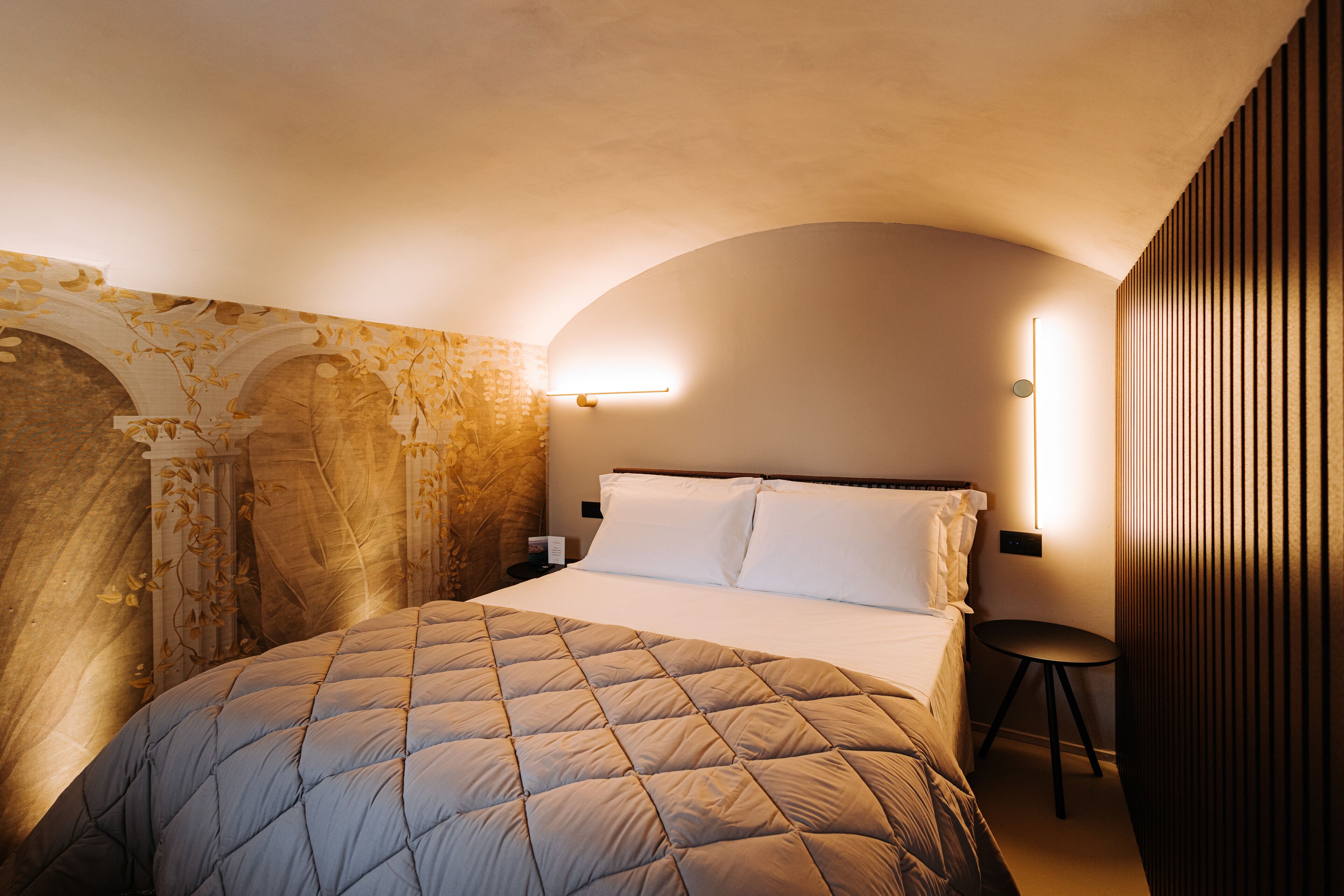 A hotel room with a bed, a small table, and a wall-mounted light fixture.