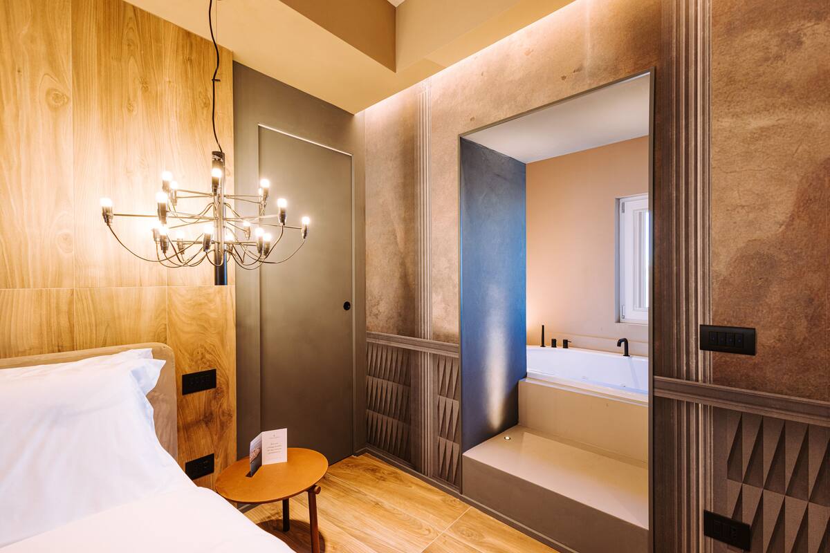 A hotel room with a wooden headboard, a chandelier, a small table, and a large mirror reflecting a bathroom with a bathtub and sink.