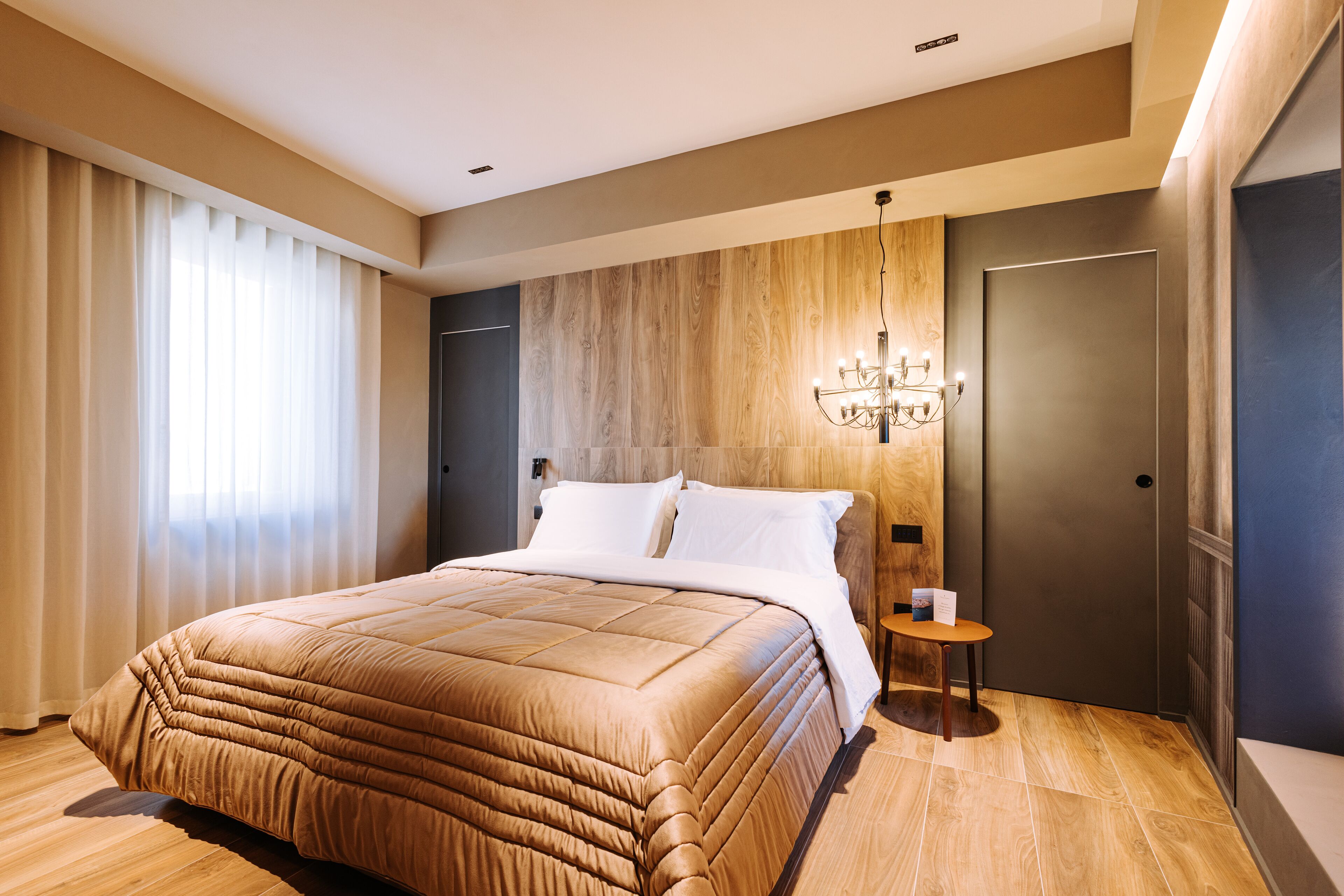 Ett modernt hotellrum med en stor säng, en vägg med träpanel, en kristallkrona och en dörr med ett handtag.