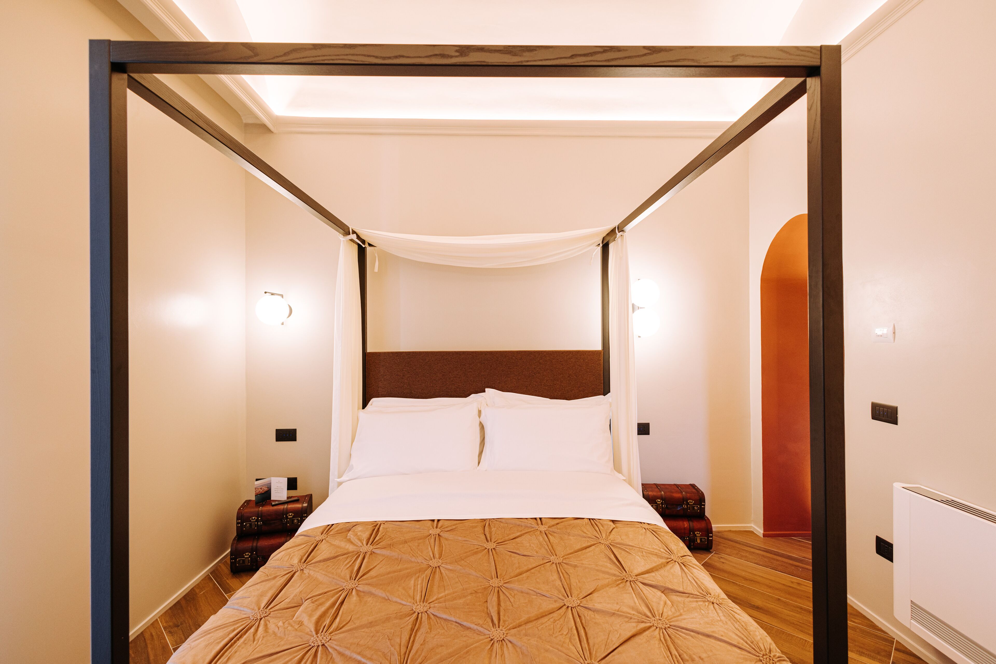 A four-poster bed with a canopy, wooden headboard, and a brown patterned bedspread. There are two bedside tables with lamps and a suitcase on one of them.