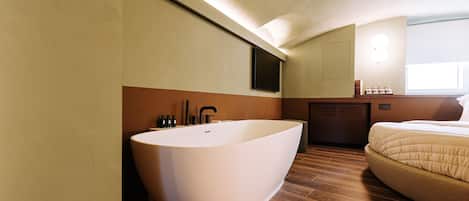 A modern bathroom with a freestanding bathtub, a wooden floor, and a bed with a beige duvet.