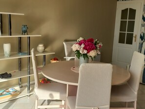 Un coin repas avec une table ronde, des chaises blanches, un vase de fleurs et des étagères remplies de divers objets.
