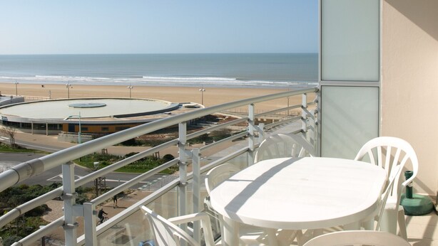 A balcony with a view of the beach and ocean. There is a roundabout with a central island, and a white table and chair set on the balcony.