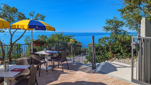 An outdoor patio with wicker furniture, a table, and a blue and yellow umbrella. - Residence Mare Azzurro (Duino-Aurisina)