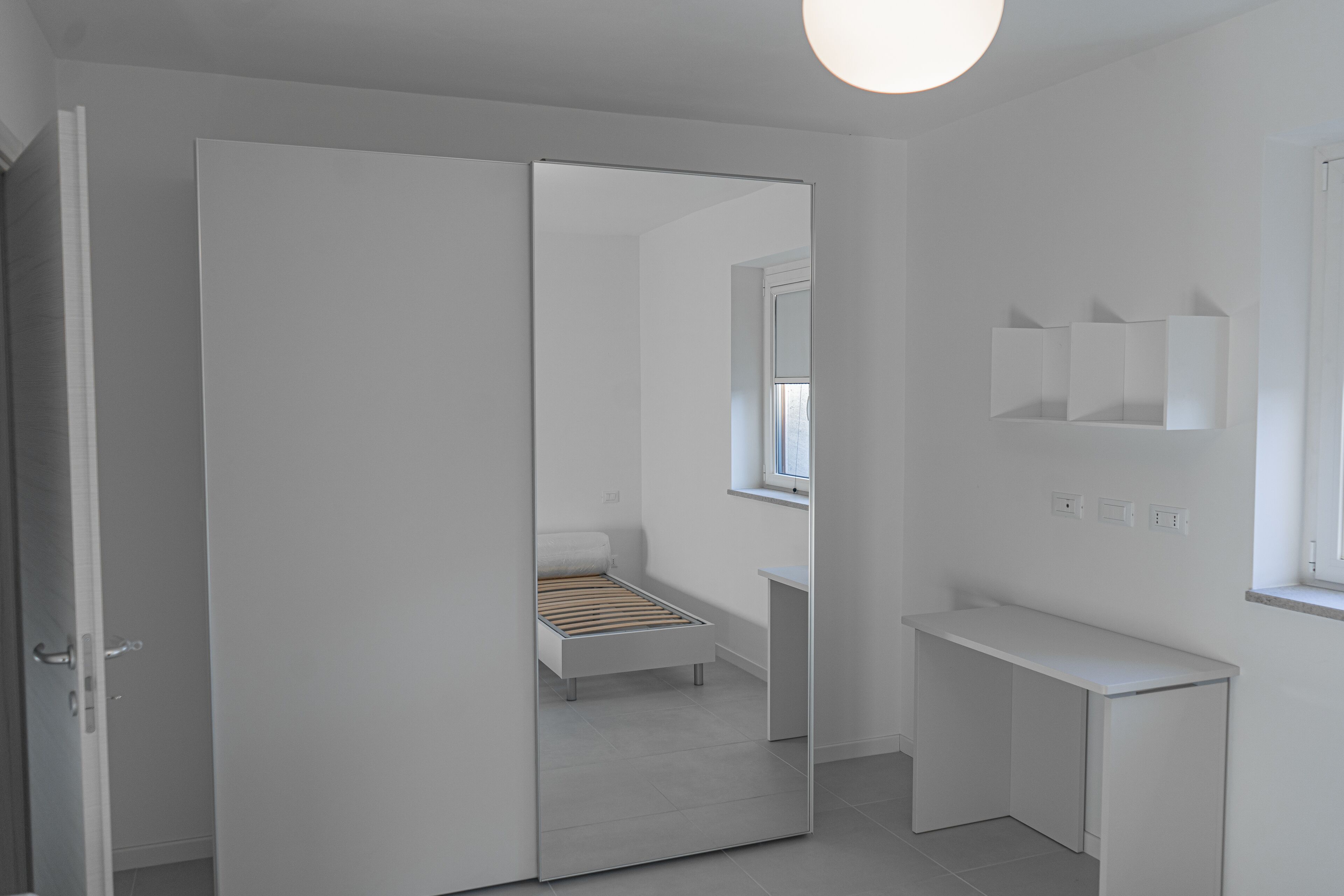 A modern, minimalist bedroom with a sliding closet door, a single bed, a bedside table, and a small window.