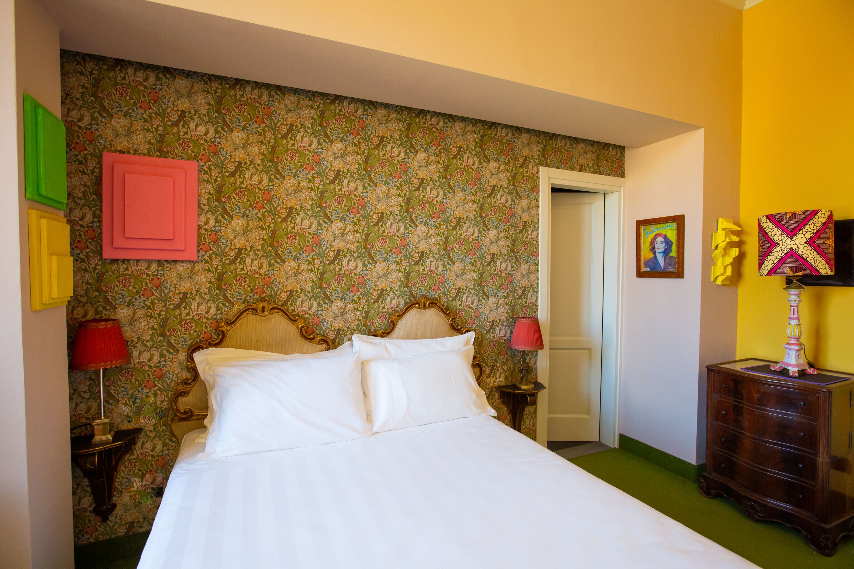 Un dormitori amb paper pintat floral, un llit amb roba de llit blanca, una nitzeta de fusta amb una làmpada i un moble armari amb una làmpada decorativa.