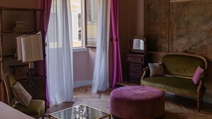 A room with a green armchair, a small round ottoman, a glass-top table with items, a wooden cabinet, and a window with curtains. - Stella d'Italia (Florence)