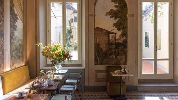 A dining area with a long table, chairs, and a vase of flowers. There are windows with views of the outside. - Stella d'Italia (Florence)