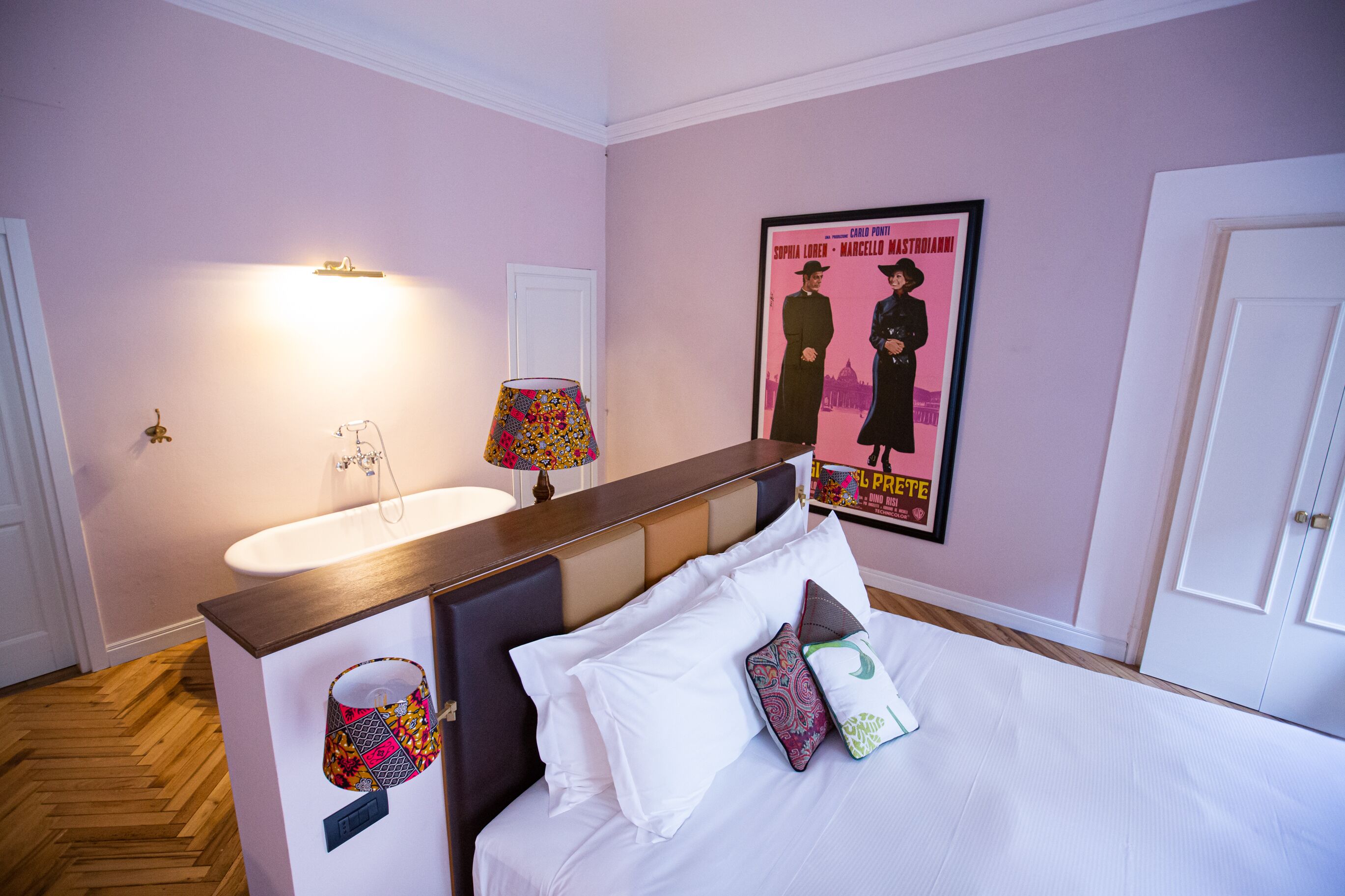 Una habitación de hotel con una cama, una mesita de noche con lámpara, un cartel en la pared y una puerta.